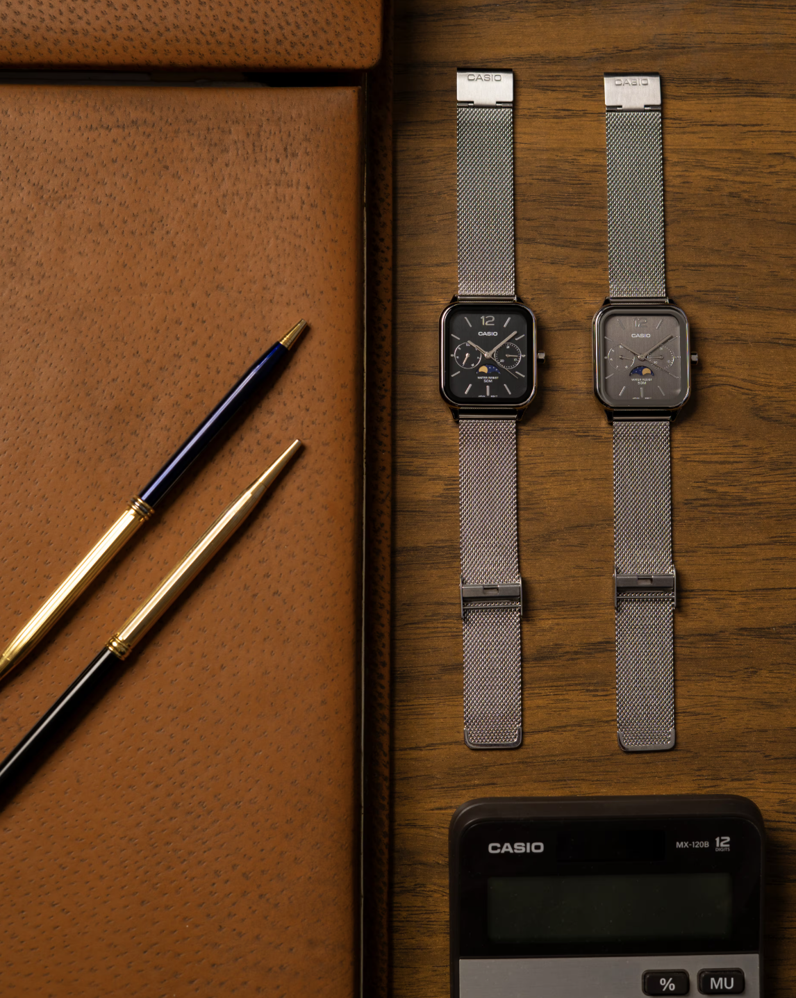 Two square silver metal watches sit on a table next to a leather pad